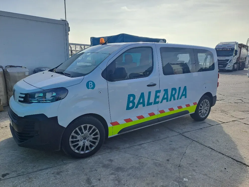 Furgoneta Balearia de color blanco con texto azul, estacionada al aire libre; franjas de seguridad amarillas y rojas.