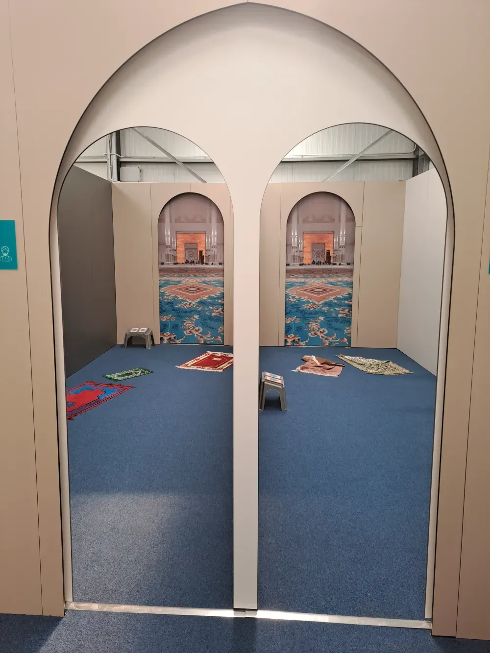 Un área de oración interior con alfombra azul y puertas arqueadas. Se ven alfombras de oración y arte mural decorativo.