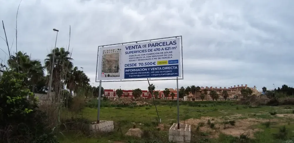 Un cartel inmobiliario que anuncia un terreno en venta frente a una nueva construcción en un día nublado.
