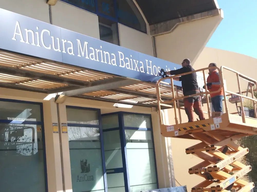 Dos personas instalando el rótulo del Hospital AniCura Marina Baixa, trabajando desde un ascensor, al aire libre.