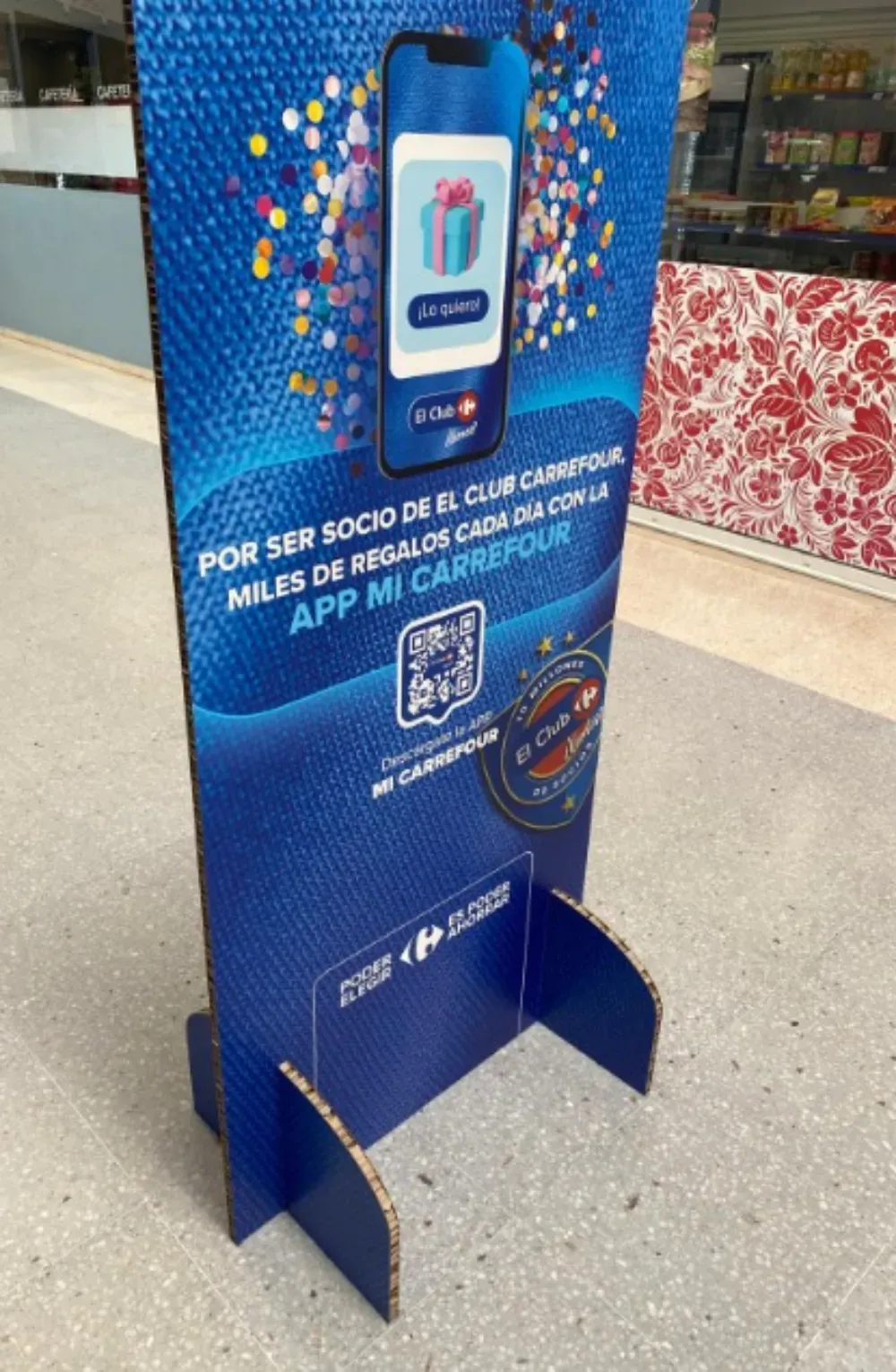 Anuncio de Carrefour en cartón con ilustración de aplicación y texto promocional en un entorno de centro comercial.
