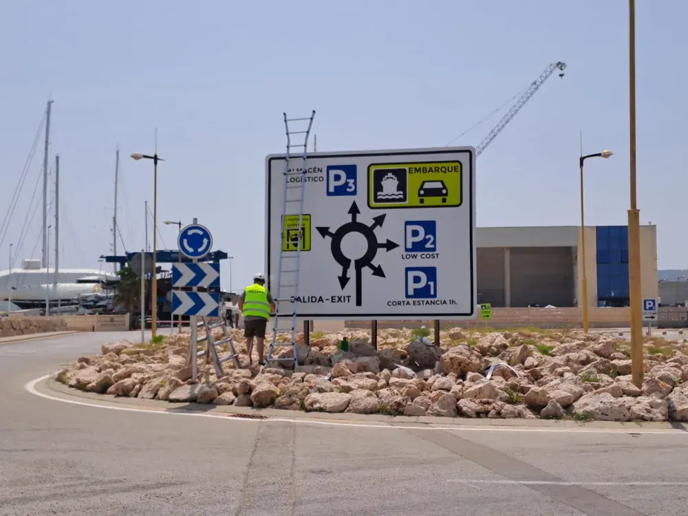 Señal en una rotonda que indica estacionamiento (P1, P2, P3), embarque y salida. Un trabajador sube a una escalera.