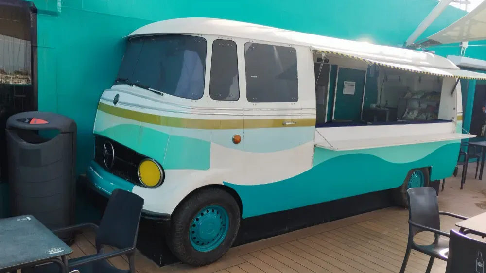 Un camión de comida blanco y verde azulado estacionado afuera, con un toldo a rayas, mesas y sillas.