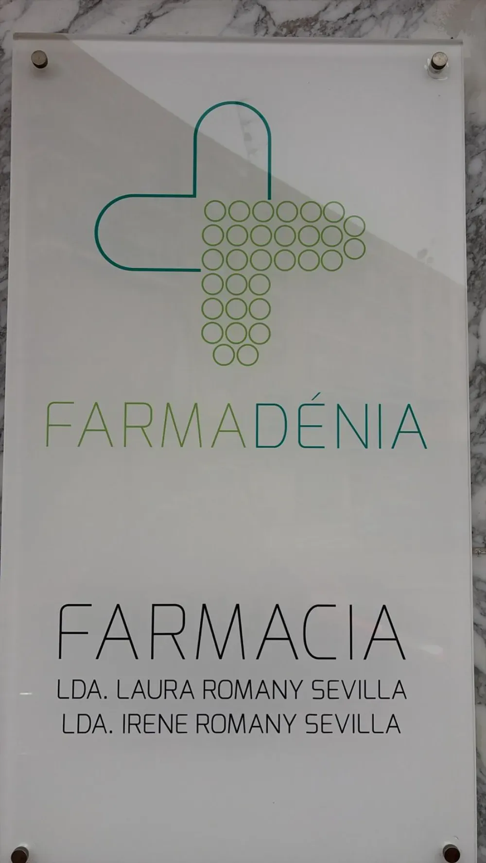 Letrero de la farmacia Farmadénia. Logotipo de la cruz verde, nombre y nombres de los farmacéuticos sobre fondo blanco.