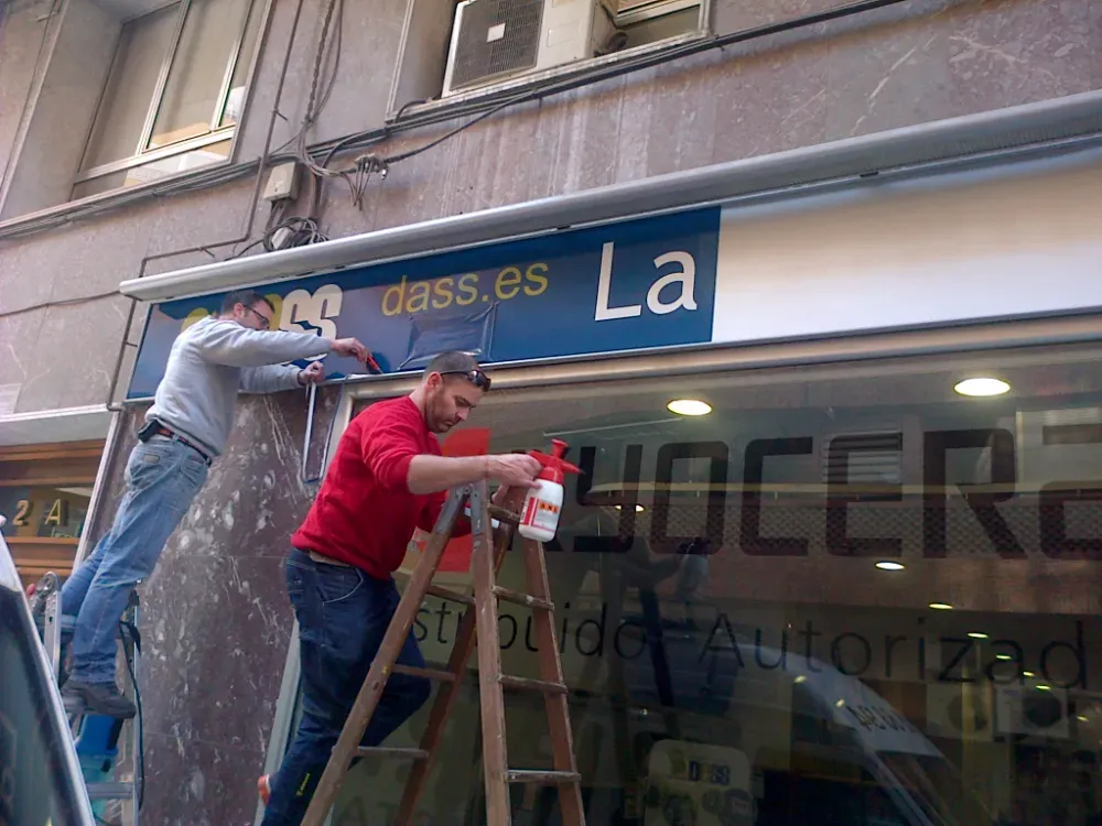 Dos personas instalando señalización encima de una tienda: una en una escalera y la otra en una plataforma.