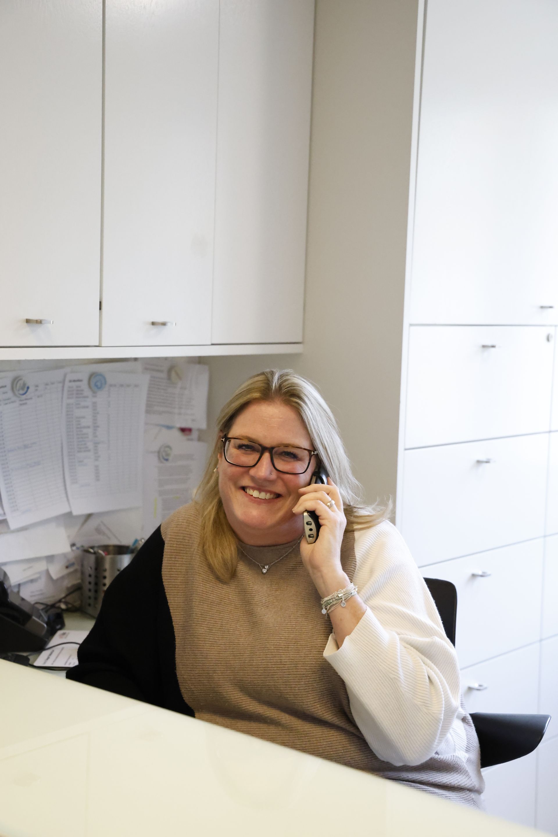 Frau mit Brille lächelt beim Telefonieren; sie sitzt an einem weißen Schreibtisch in einem Büro.