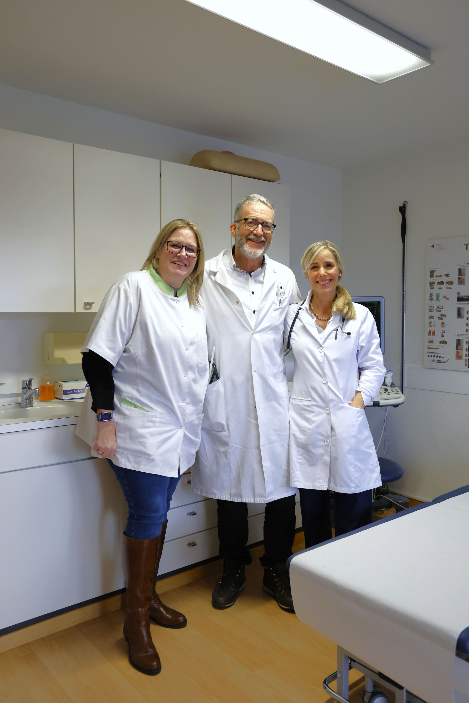 Drei medizinische Fachkräfte stehen in einem Büro. Eine trägt einen Laborkittel, zwei weiße Hemden, eine Jeans und Stiefel.