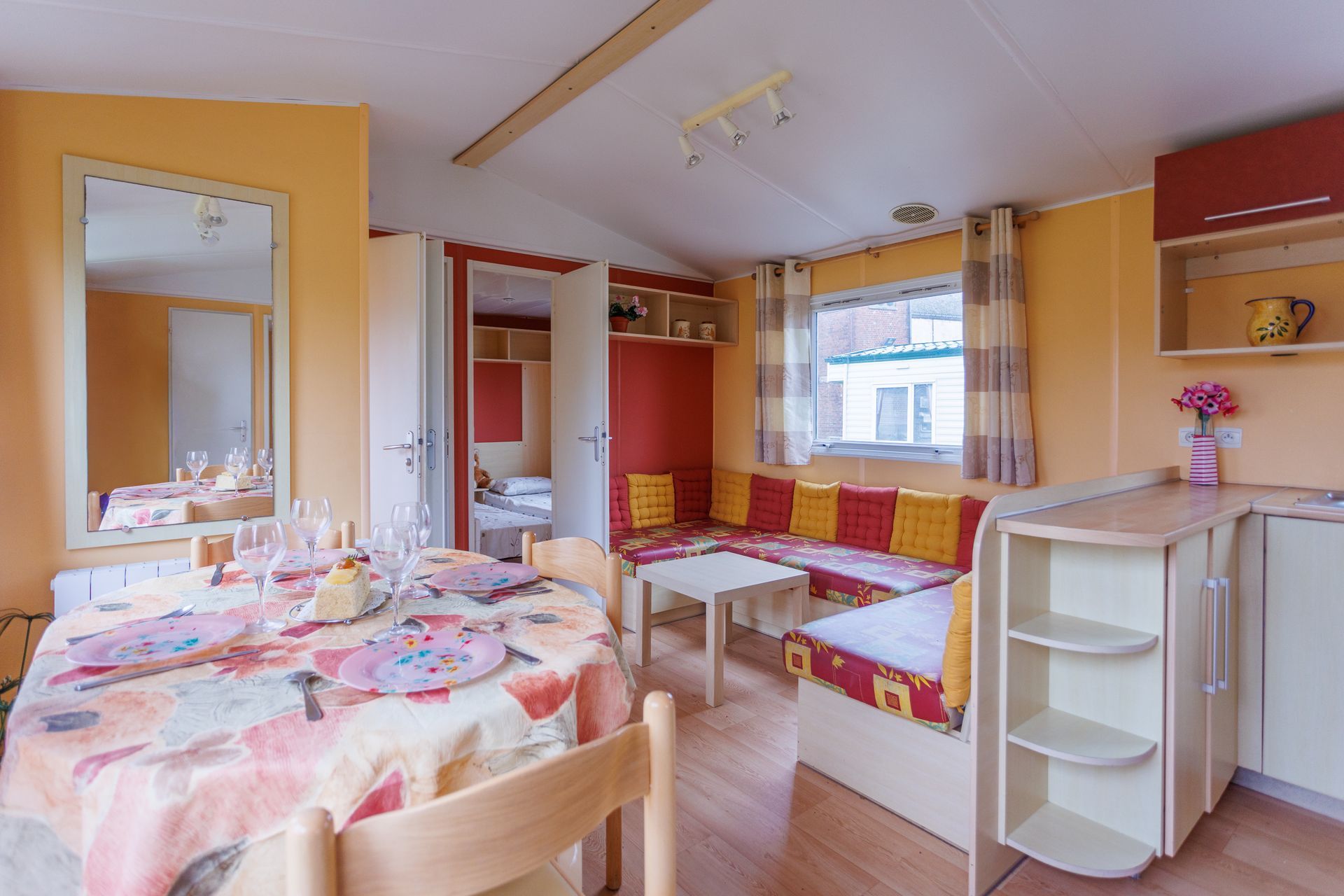 Intérieur d'une maison mobile : coin repas, banquette intégrée, cuisine et porte donnant sur une chambre.
