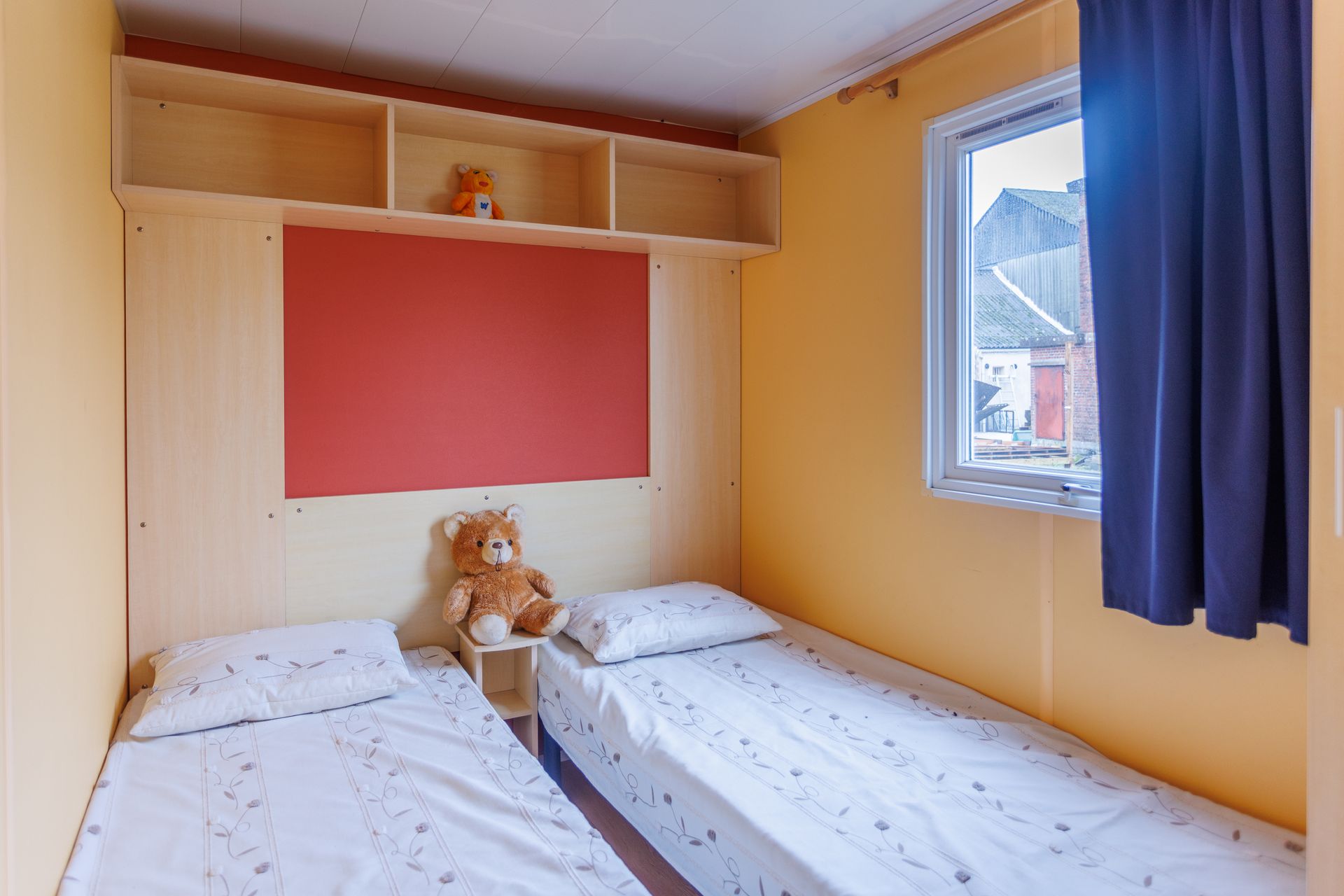Deux lits jumeaux dans une chambre jaune avec un mur d'accent rouge, des étagères intégrées et une fenêtre avec un rideau bleu.