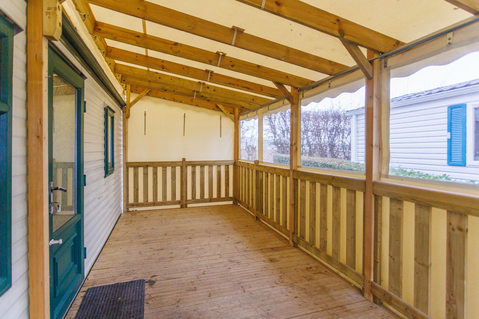 Véranda couverte avec balustrade en bois, plafond et toile, jouxtant un bâtiment blanc.