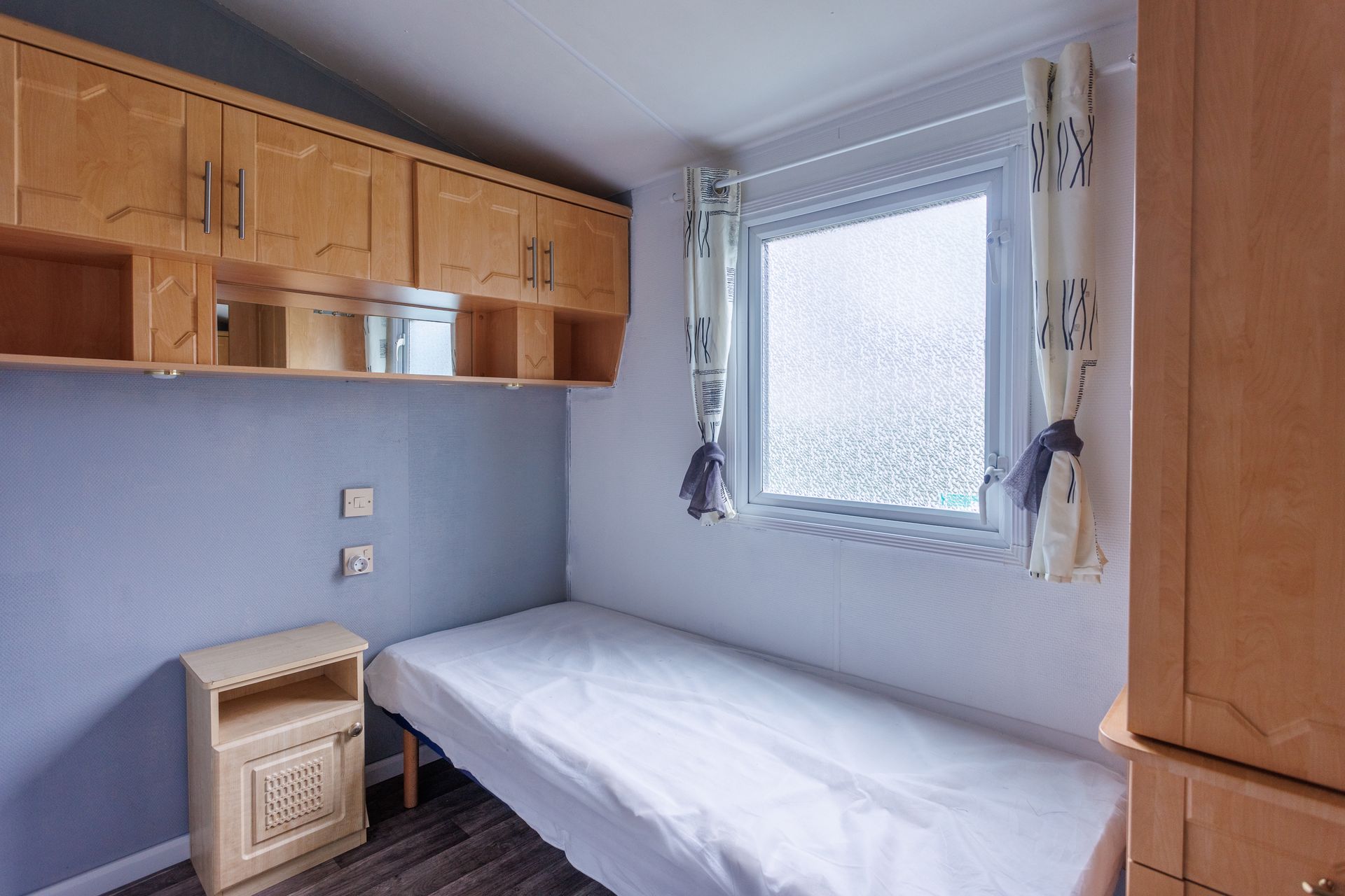 Une petite chambre avec un lit simple, une table de chevet, des armoires en bois et une fenêtre.