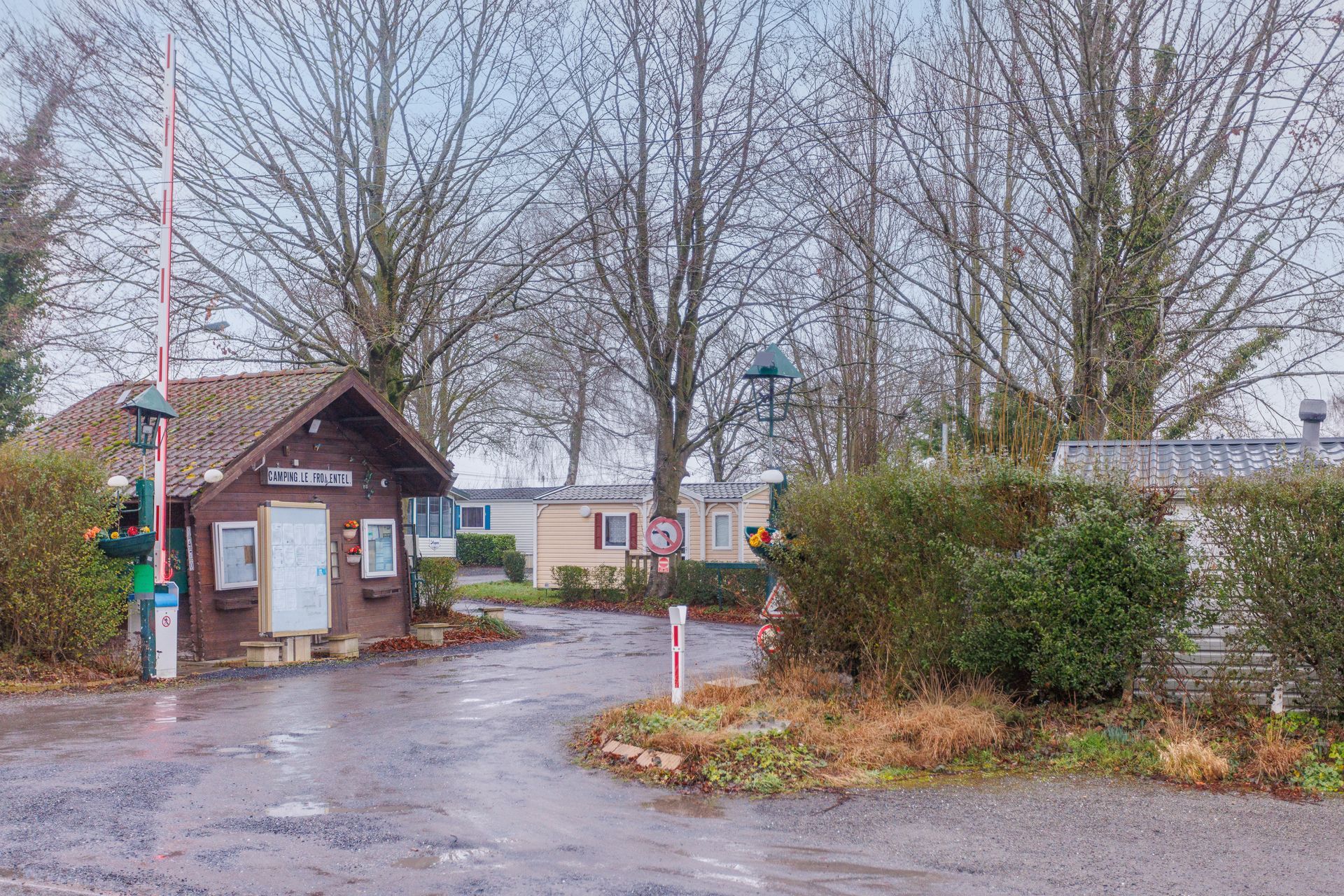 Entrée d'un terrain de camping avec un poste de garde, un portail et des arbres dénudés sous un ciel couvert.