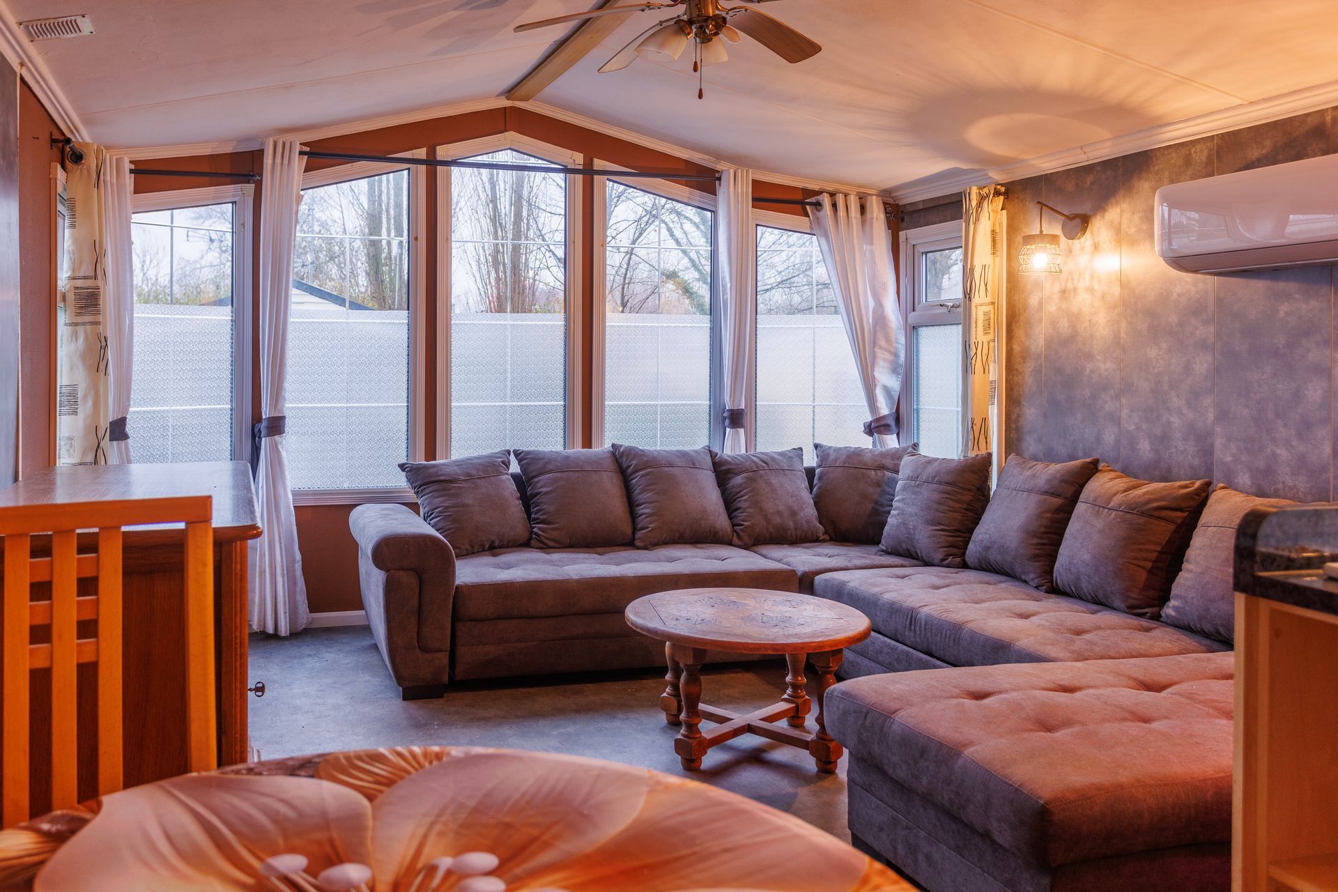 Salon avec canapé d'angle, table basse et grandes fenêtres. Tons bruns et gris.