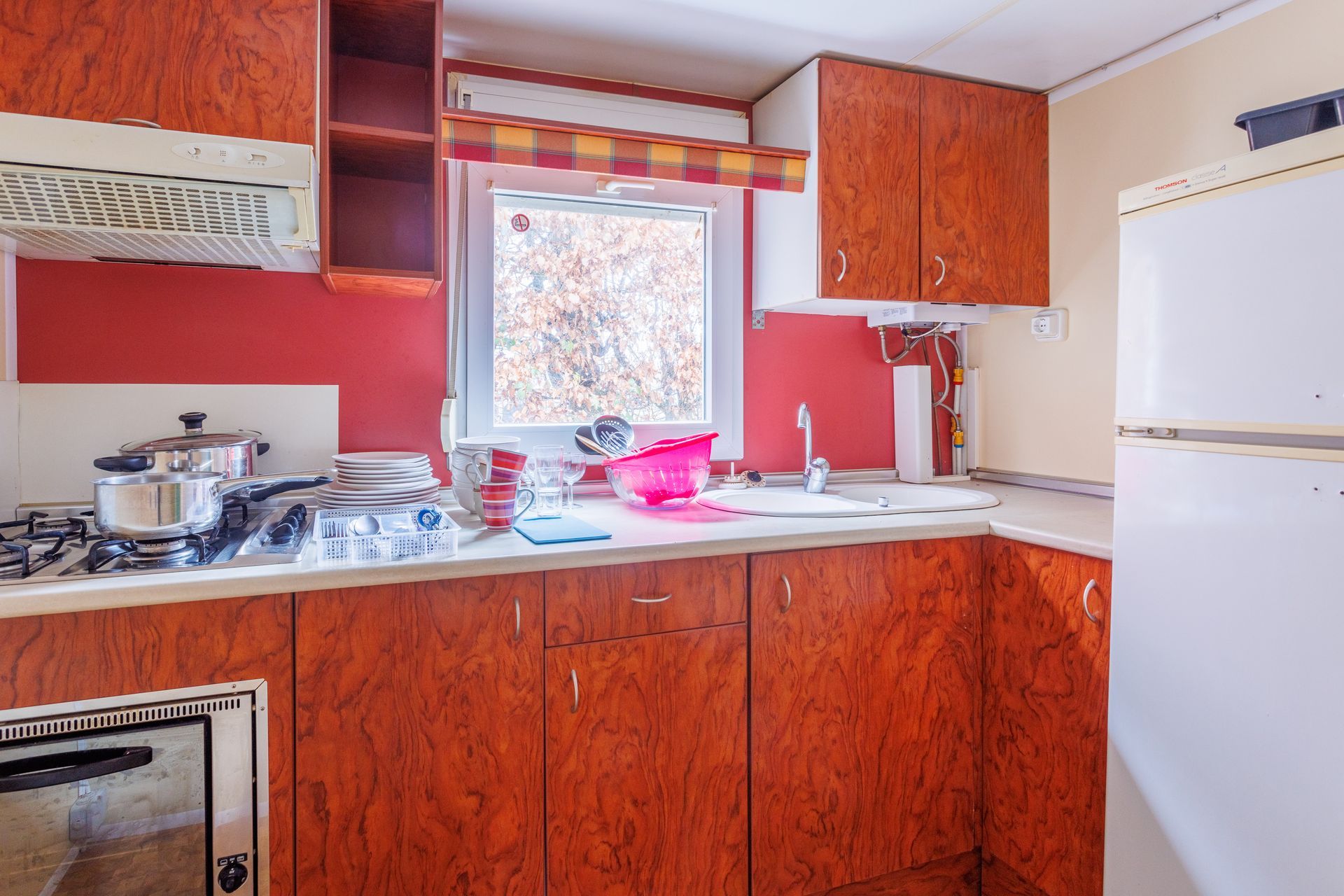 Cuisine avec placards rougeâtres, crédence rouge, cuisinière, évier, fenêtre et réfrigérateur.