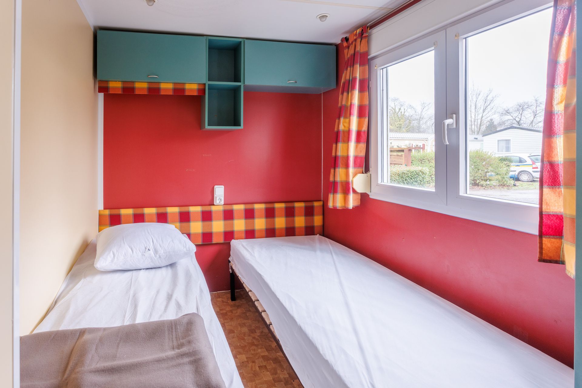 Deux lits dans une petite chambre aux murs rouges, avec fenêtre et rangements.