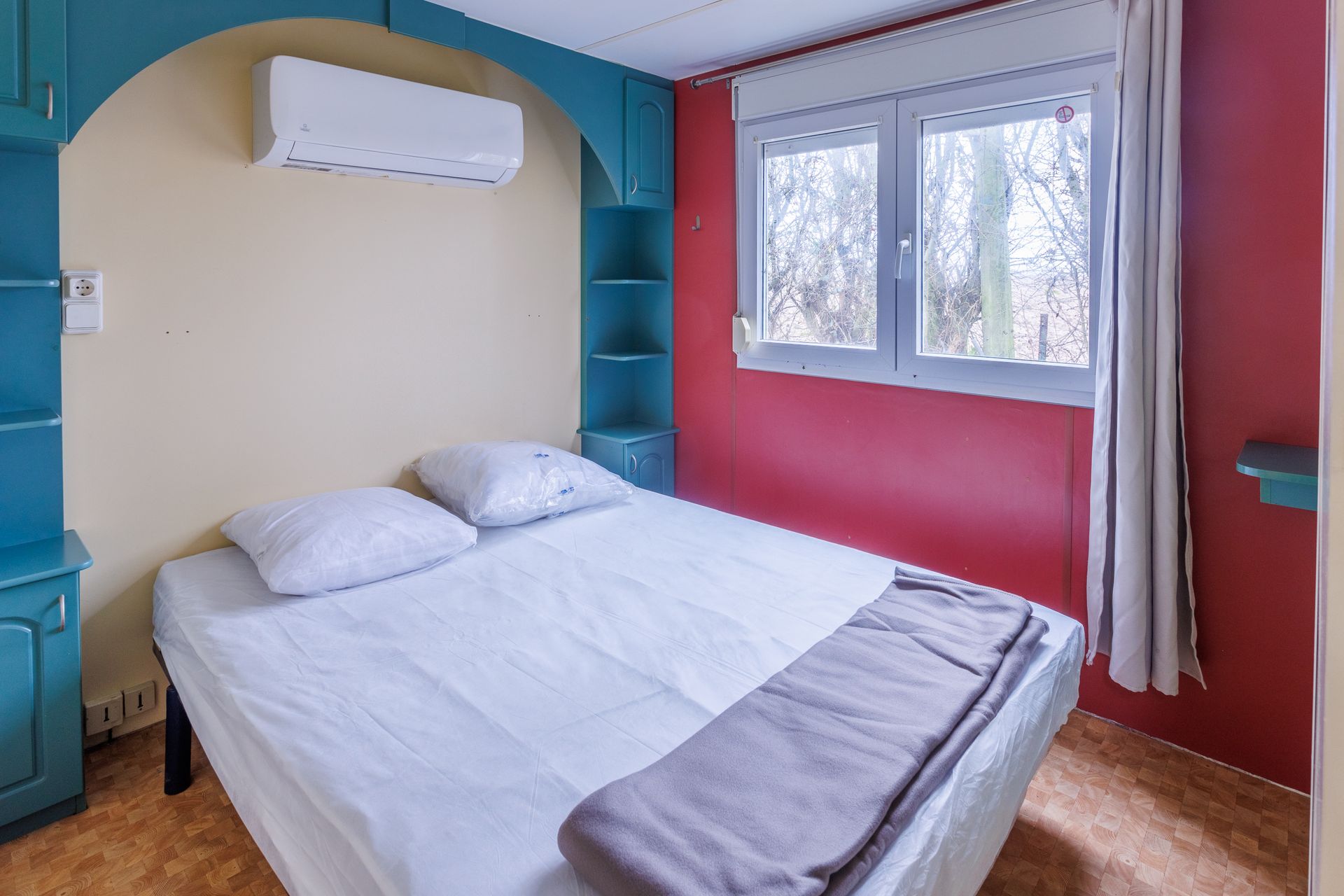 Chambre avec un lit, des étagères de rangement bleues et une fenêtre donnant sur les arbres.