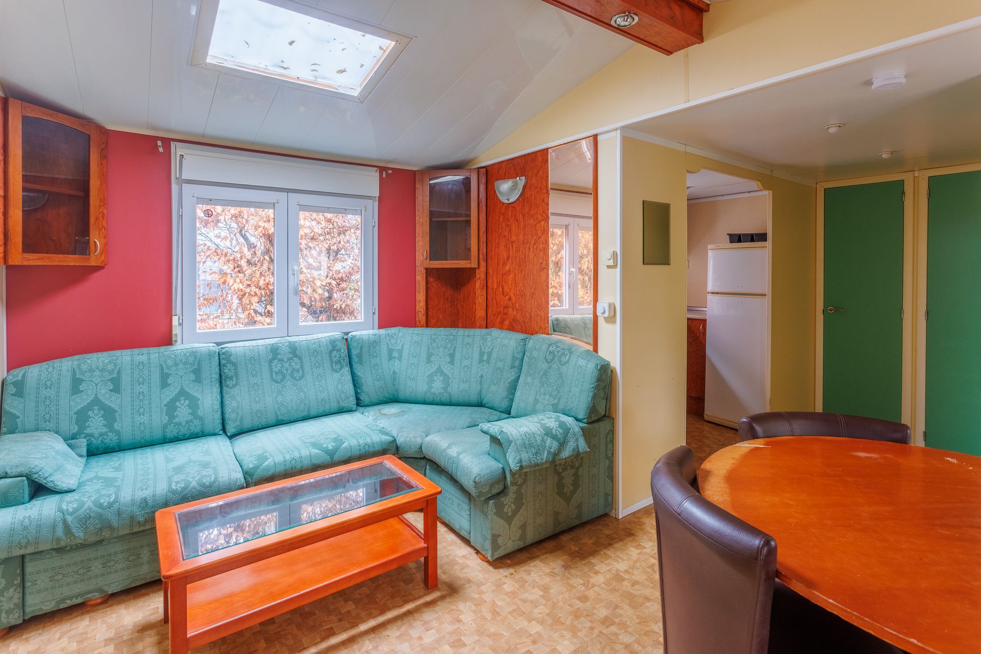 Salon avec canapé turquoise, mur rouge, table orange et vue sur la cuisine.