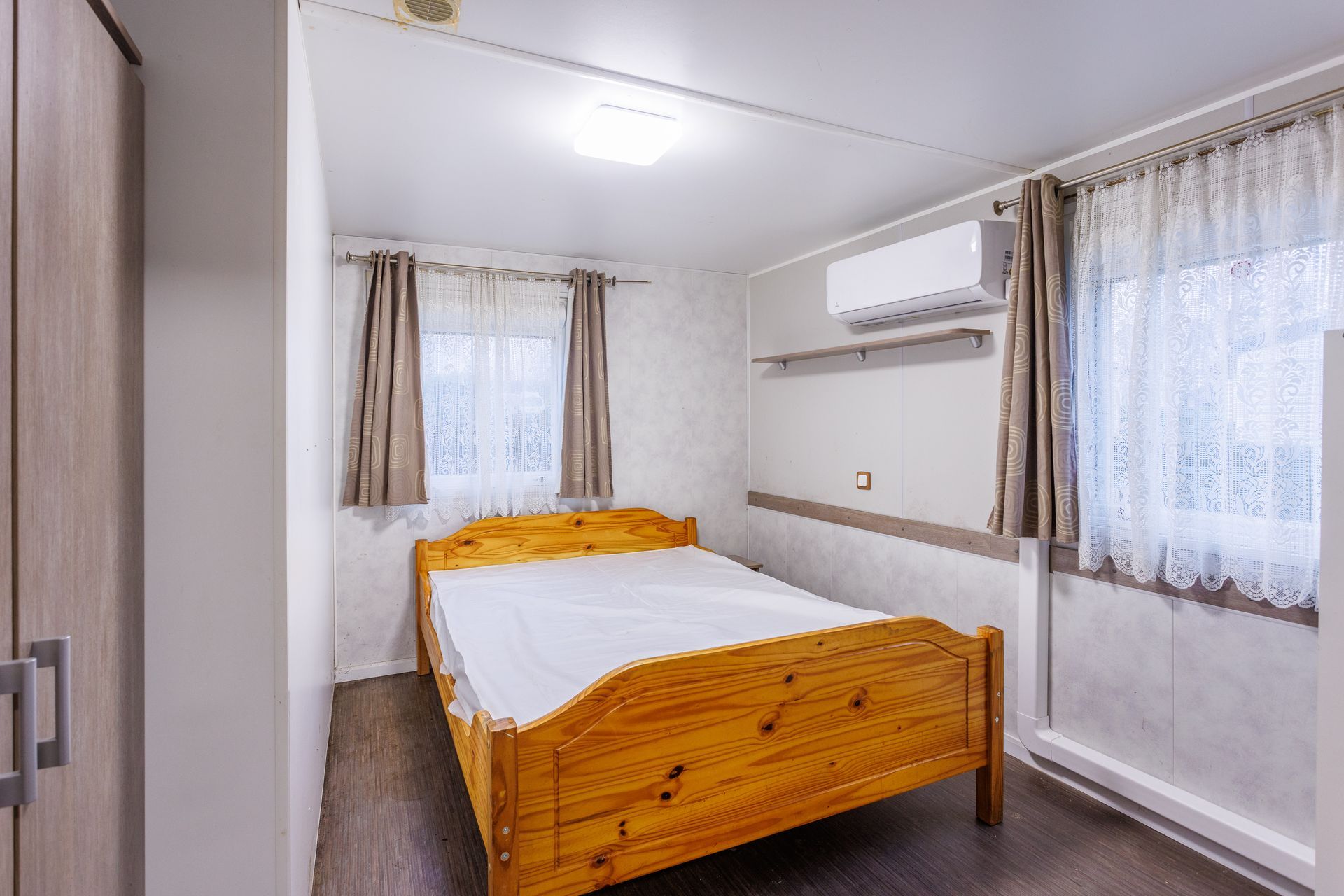 Intérieur de chambre avec un lit en bois, deux fenêtres avec rideaux et un climatiseur.