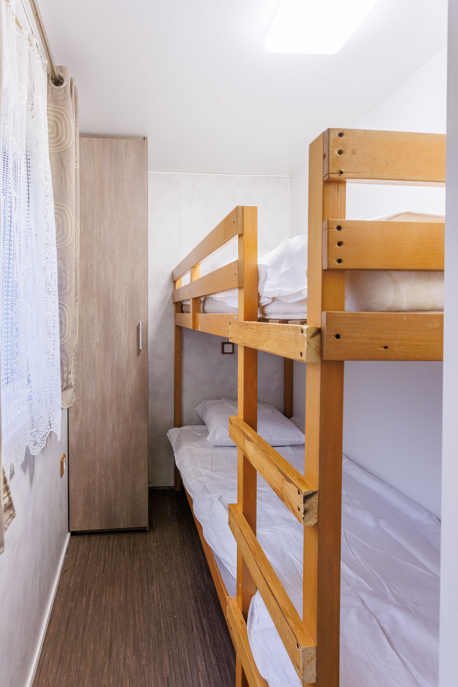 Lits superposés dans une petite chambre avec placard et fenêtre. Linge de lit blanc, meubles en bois clair et murs blancs.