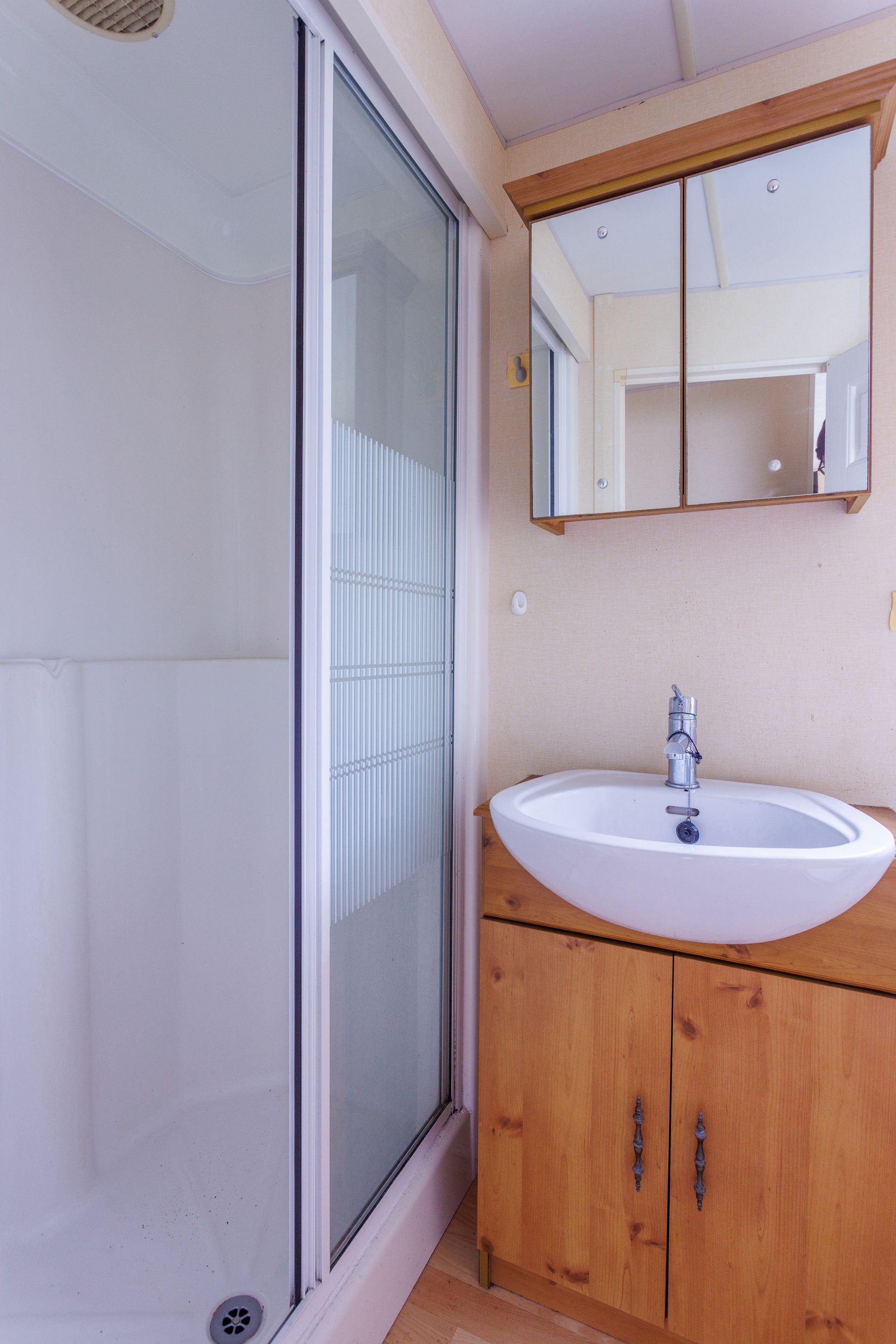 Salle de bains avec douche, meuble-lavabo et armoire à pharmacie avec miroir. Murs beiges, meubles en bois clair.