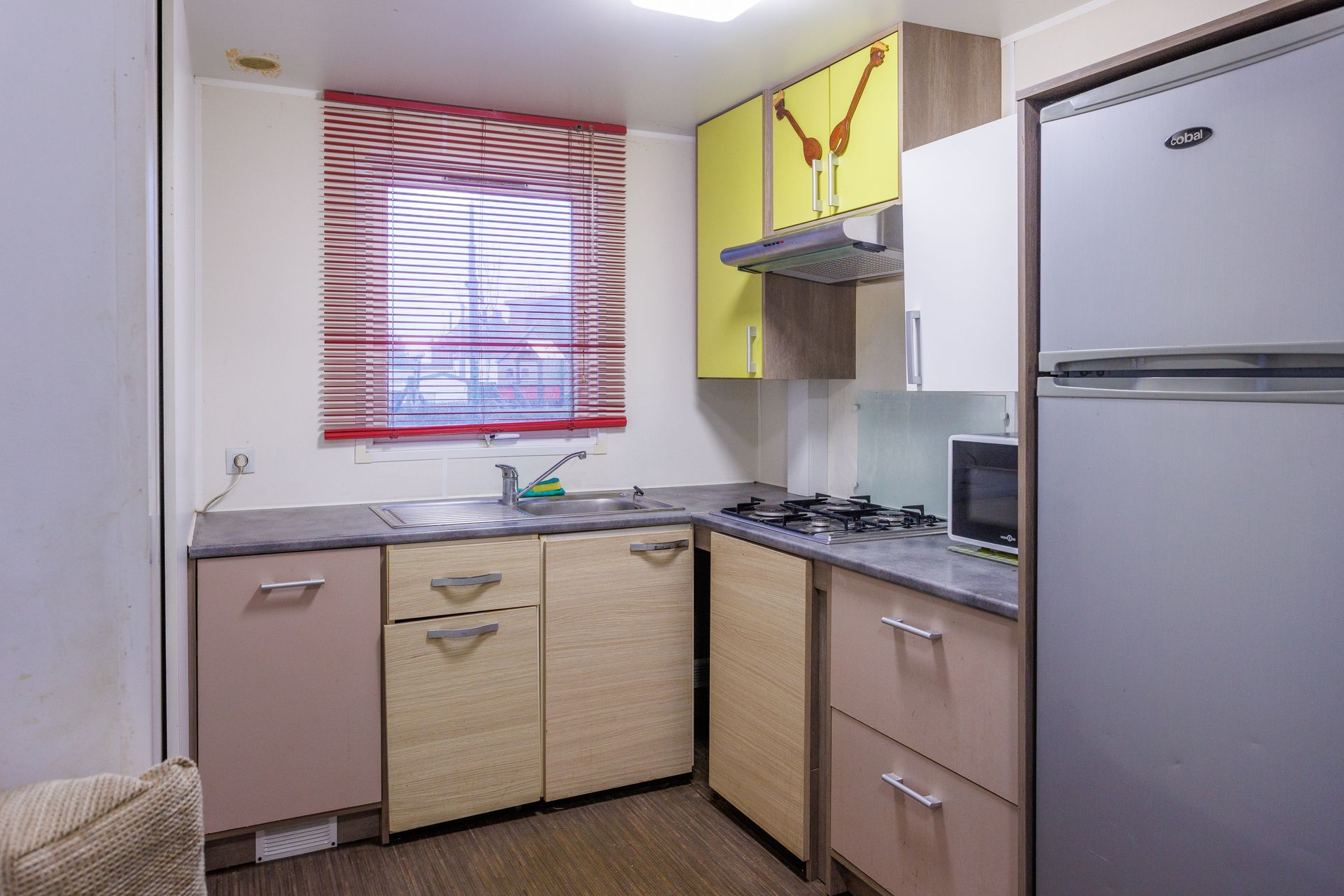 Petite cuisine avec placards, cuisinière, évier, réfrigérateur et une fenêtre avec stores rouges.