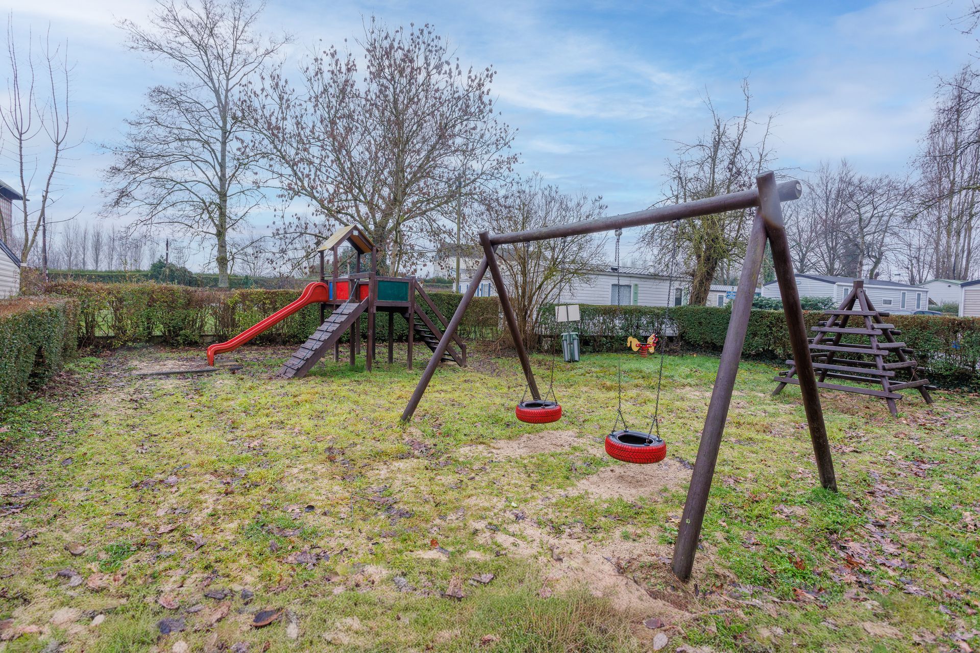 Aire de jeux avec balançoires, toboggan et structure d'escalade sur une pelouse, avec des arbres en arrière-plan.