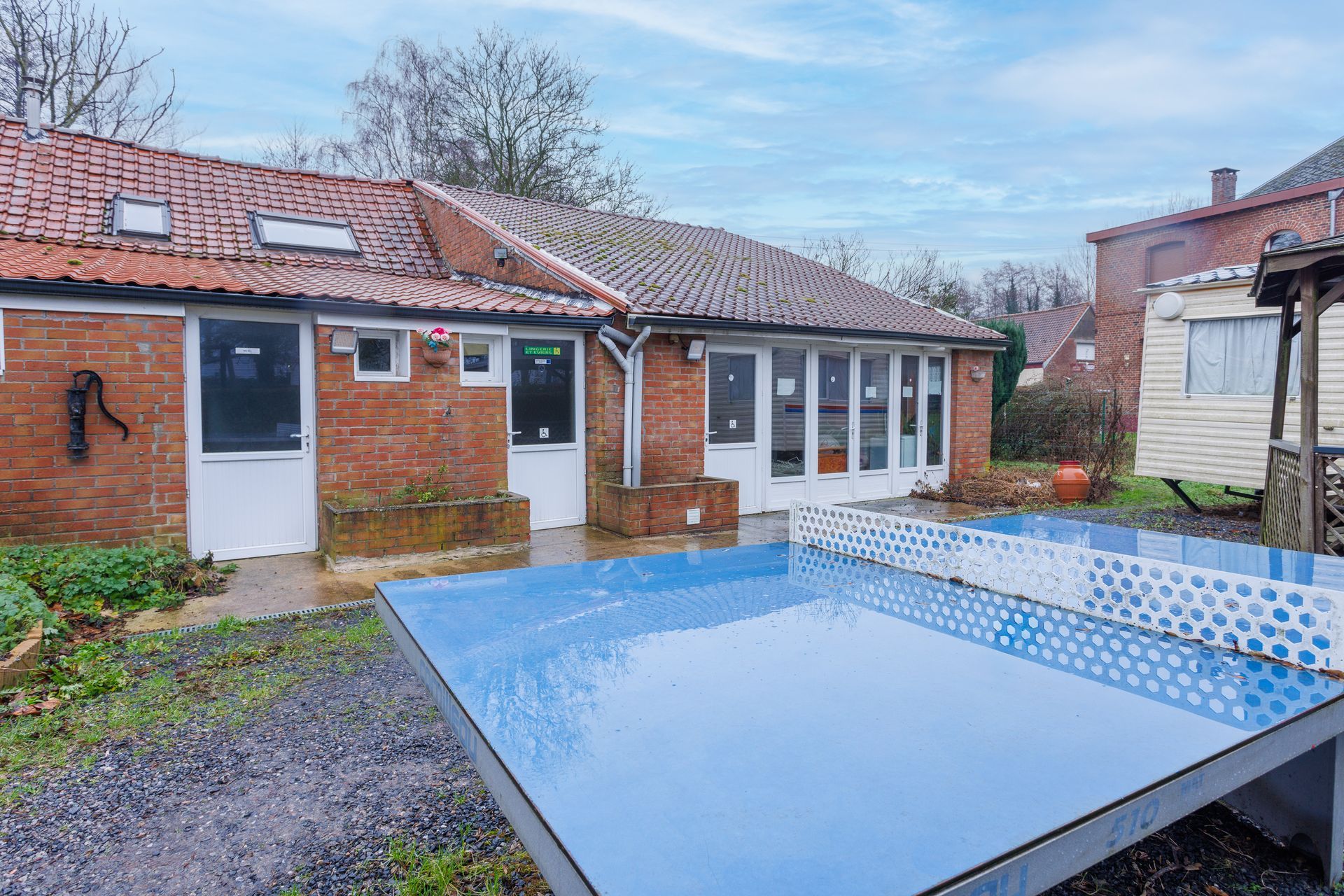 Vue extérieure avec une table de ping-pong bleue au premier plan, un bâtiment en briques et une caravane à l'arrière-plan.