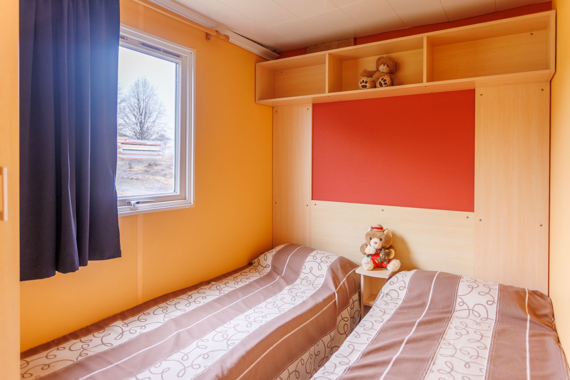 Deux lits jumeaux dans une chambre aux murs orange, une fenêtre avec un rideau foncé et des étagères intégrées.