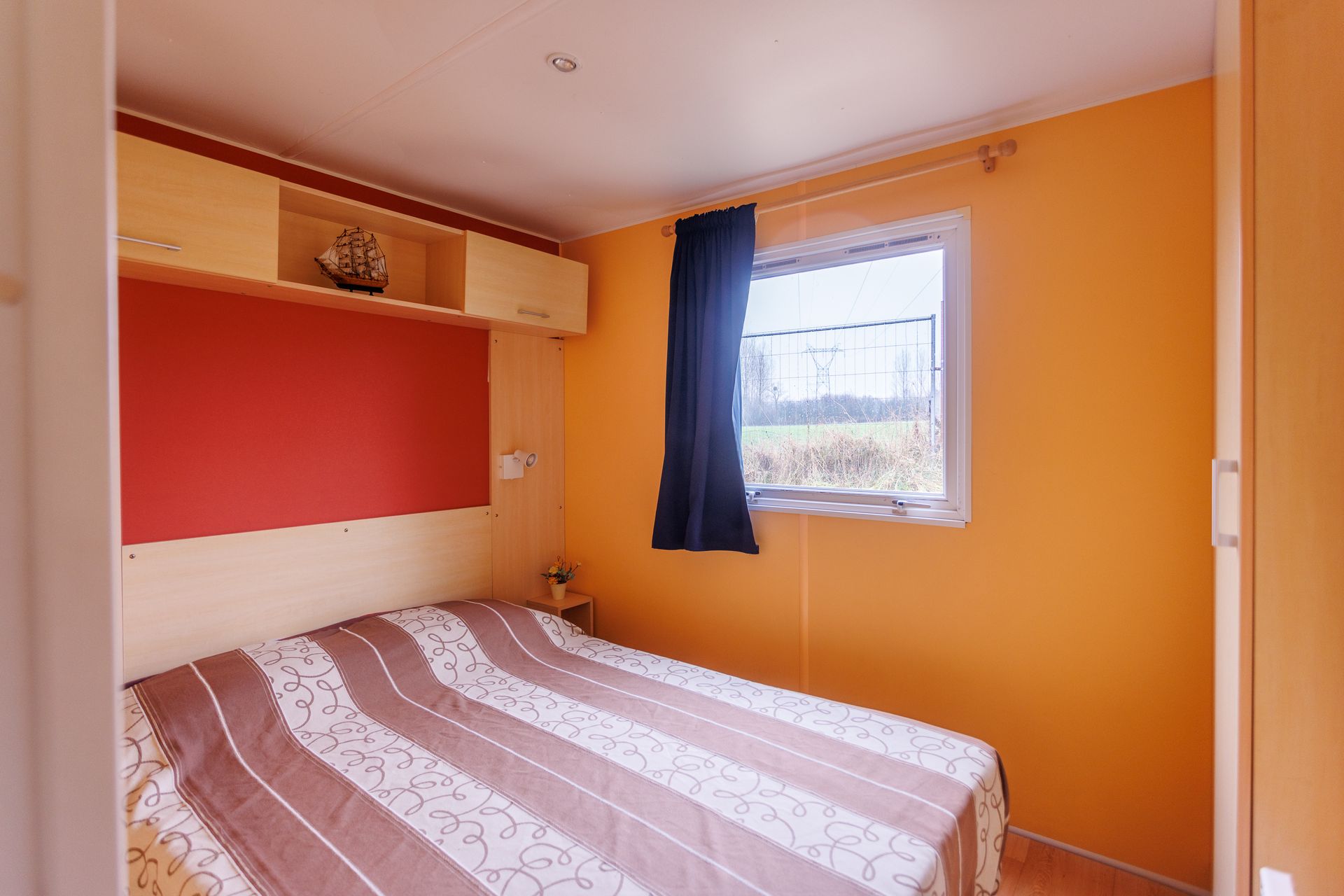Chambre avec lit, murs orange, fenêtre avec rideau foncé, rangements intégrés.