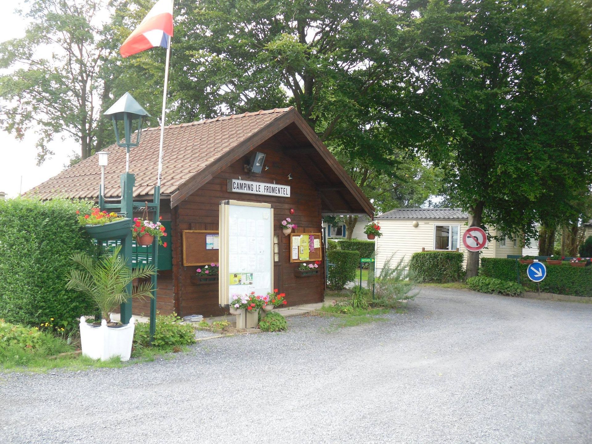 Petit bâtiment en bois, probablement le bureau du camping, avec un drapeau et des panneaux d'information. Des arbres et de la verdure entourent le bâtiment.