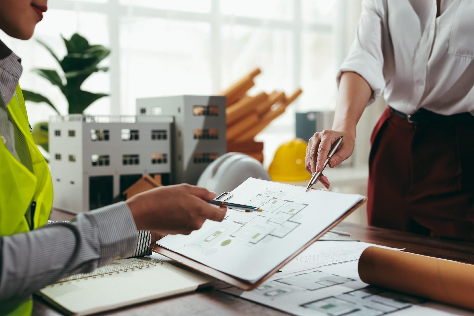 Zwei Personen prüfen an einem Schreibtisch Architekturpläne, in der Nähe stehen Modelle und Baupläne.
