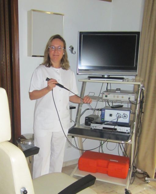 Una mujer con bata blanca sostiene un endoscopio en un consultorio médico, de pie junto a un equipo con un monitor.