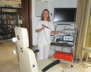 Médico con uniforme blanco sosteniendo un endoscopio, de pie cerca de un equipo médico y una silla de examen.