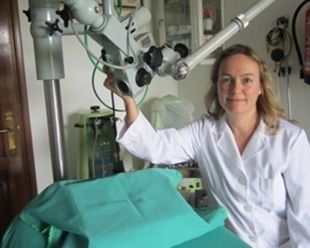 Mujer con bata blanca, sonriendo, junto a un microscopio quirúrgico en un consultorio médico.
