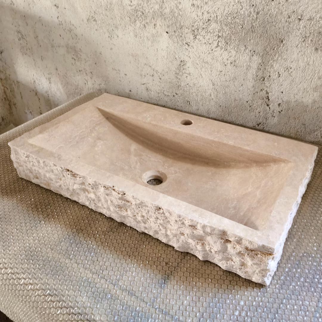 Lavabo rectangulaire en pierre beige, avec vasque sculptée et bords irréguliers, sur une surface texturée.