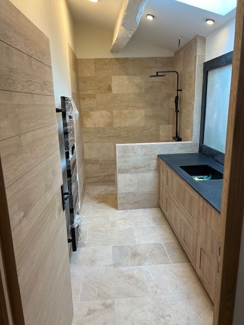 Salle de bains avec carrelage beige, meubles en chêne, robinetterie noire et plan de travail noir.