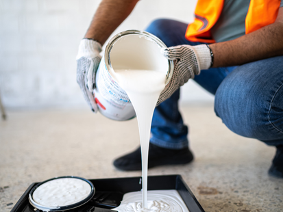 Personne versant de la peinture blanche d'un pot dans un bac à peinture, portant des gants de travail et un jean.