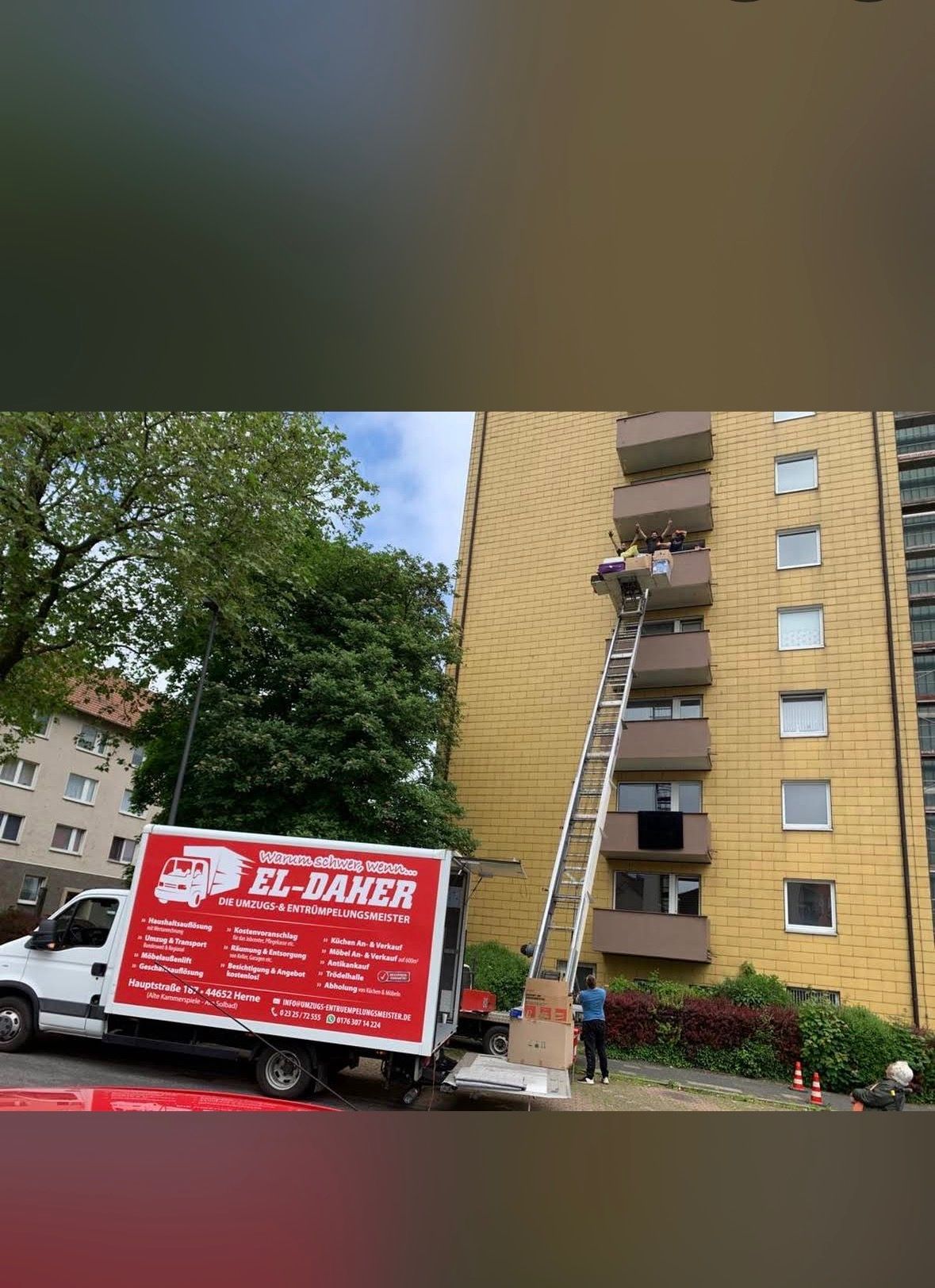 Ein Umzugswagen, der vor einem hohen gelben Wohnhaus parkt, benutzt einen Möbellift, um Möbel aus einer Wohnung zu befördern.