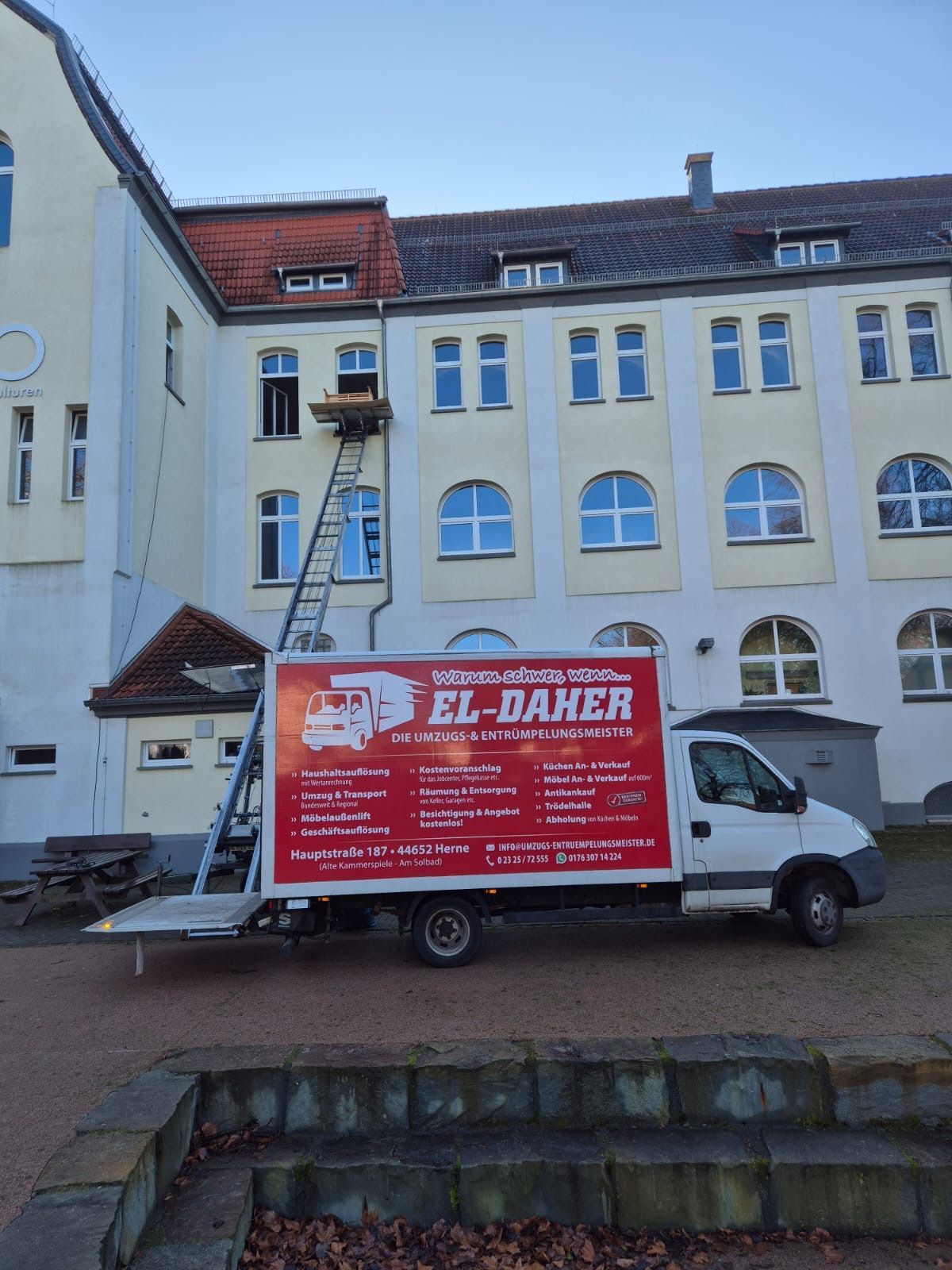 Ein weißer El-Daher-Umzugswagen mit Möbellift, der bis zu einem Fenster im zweiten Stock eines großen, hellen Gebäudes reicht.