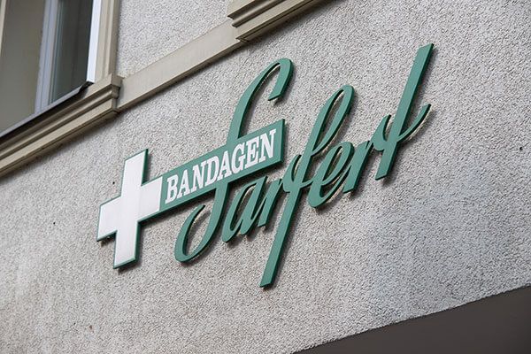 Schild für Bandagen Surfert, eine Apotheke. Weißes Kreuz und grüne Schrift auf einer Stuckwand.