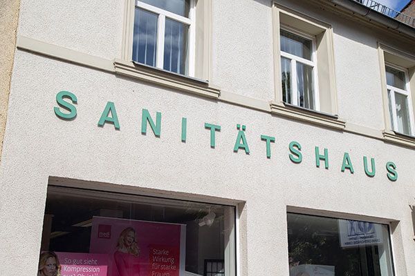 Schild „Sanitätshaus“ an einem Gebäude mit grüner Schrift über einer Ladenfront; Fenster und weiße Außenwände.“