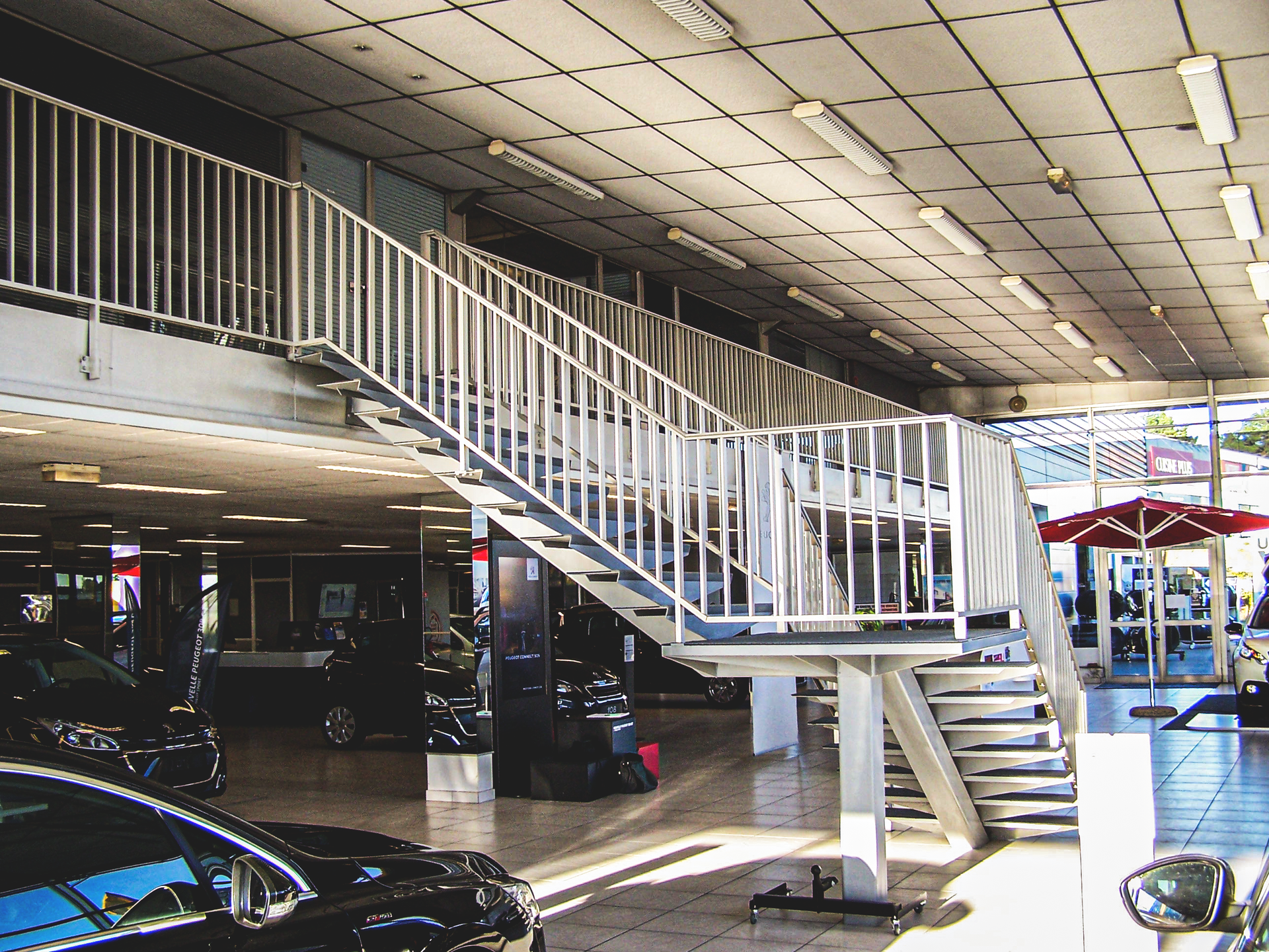 Escalier métallique dans un showroom automobile. Voitures neuves visibles au rez-de-chaussée et à l'étage. Éclairage intense.