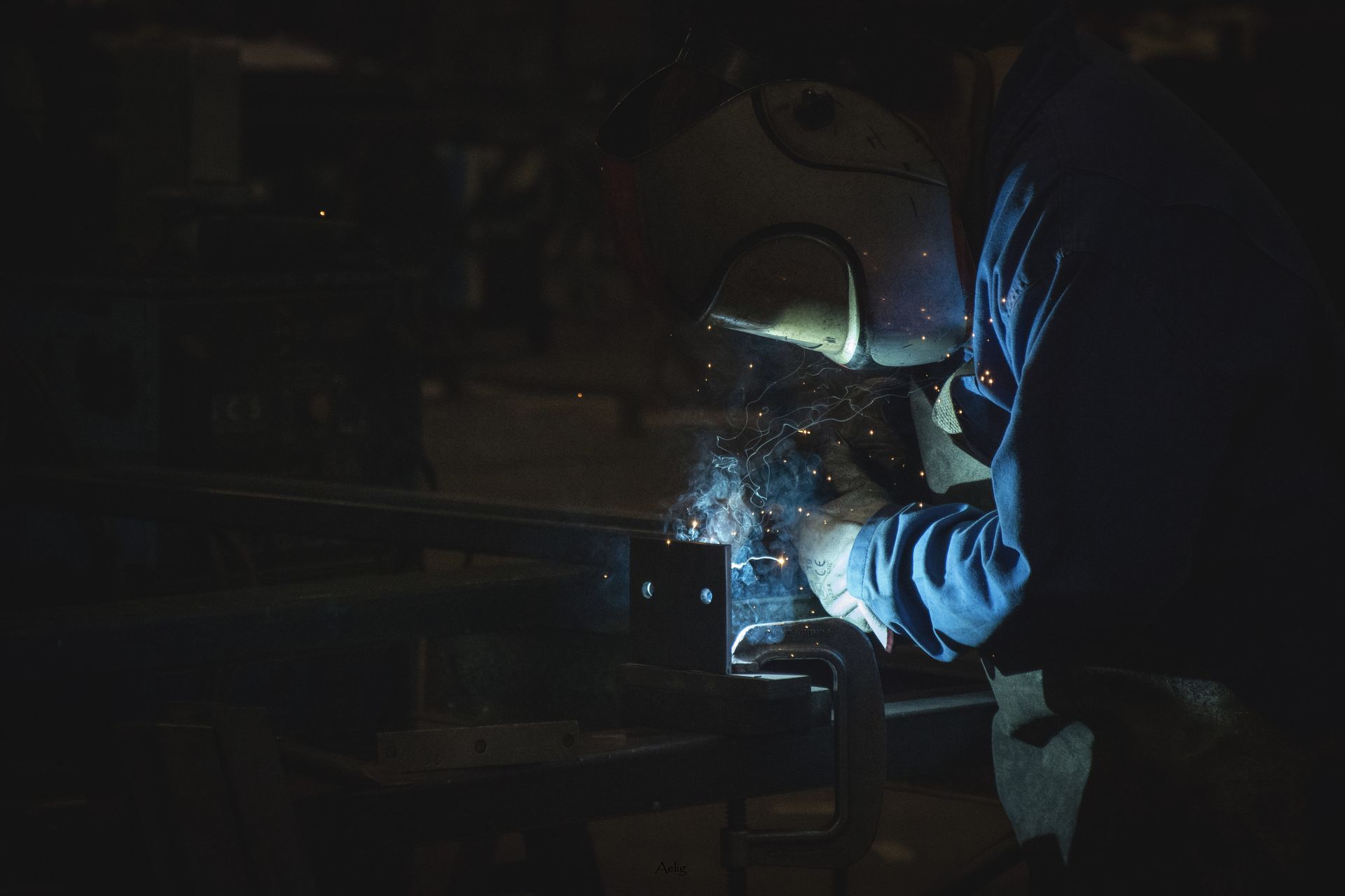 Une personne utilise une scie, des étincelles jaillissent. Environnement sombre, gants et vêtements bleus.