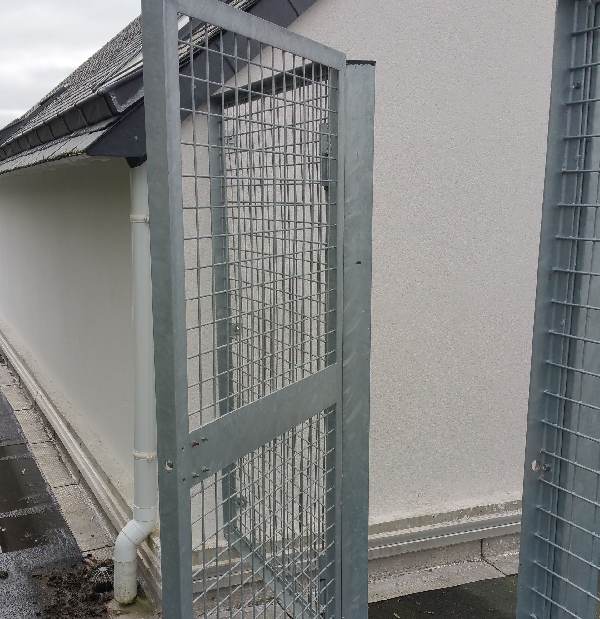 Portail métallique ouvert devant un bâtiment blanc. Toiture et gouttière grises visibles.