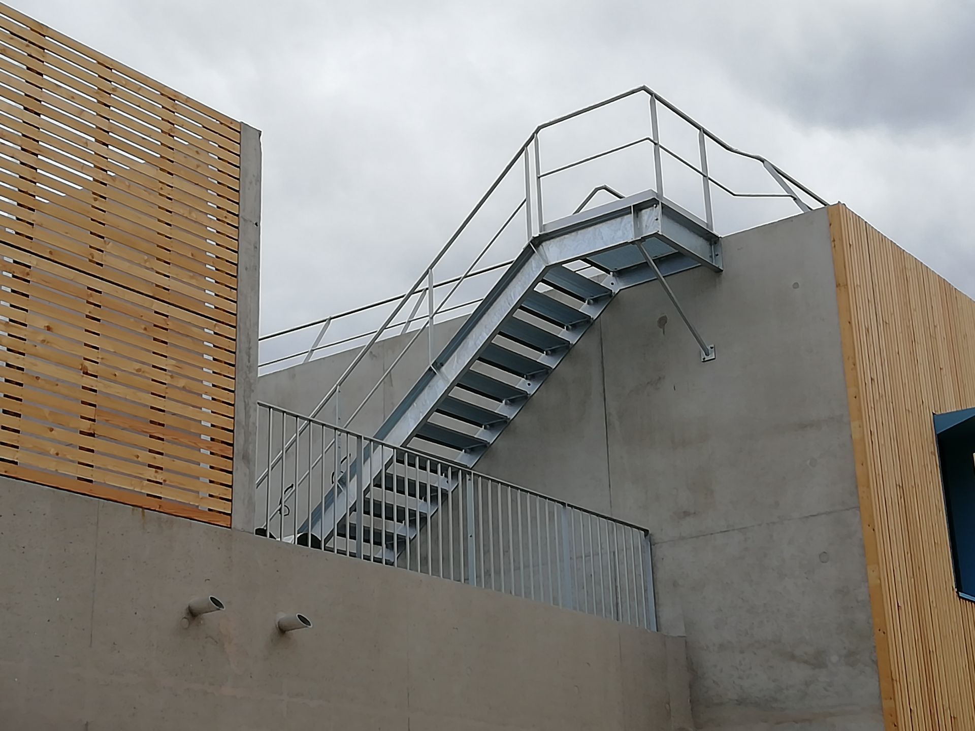 Escalier extérieur en métal sur un bâtiment avec des panneaux en bois et un ciel nuageux.