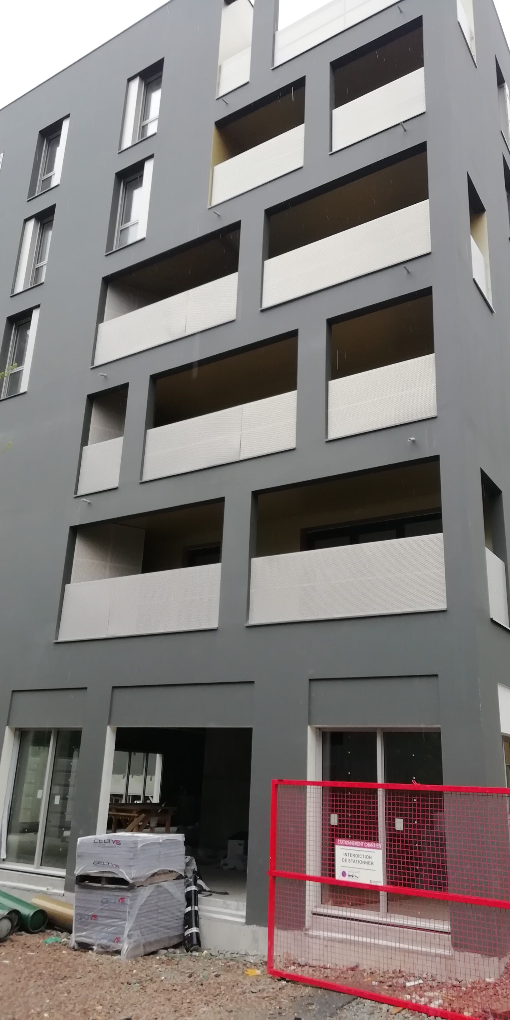 Immeuble d'appartements gris moderne avec balcons rectangulaires. Matériaux de construction et clôture rouge devant.
