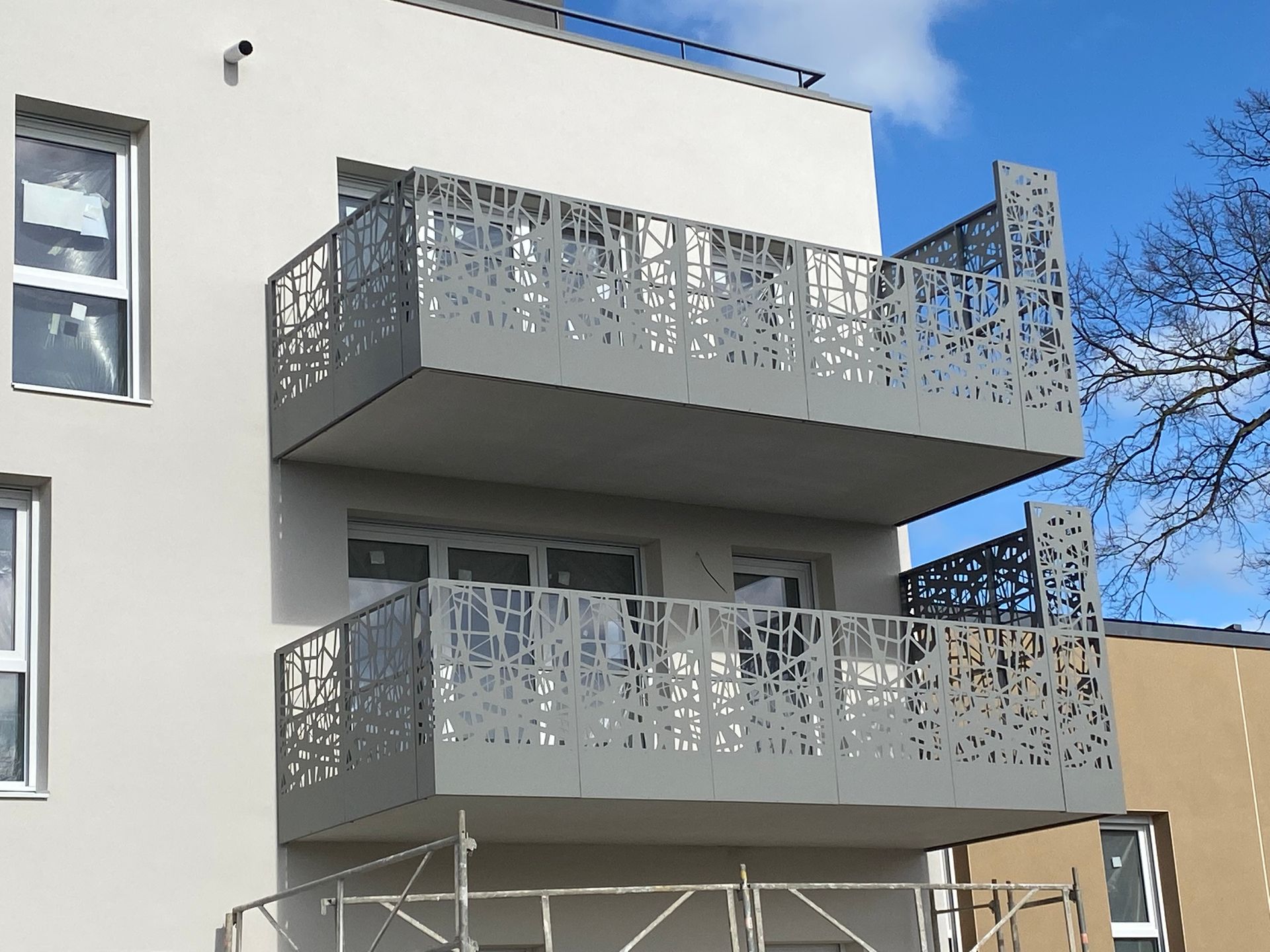 Bâtiment de deux étages avec balcons gris. Garde-corps métalliques ornés d'une découpe décorative. Extérieur gris clair.