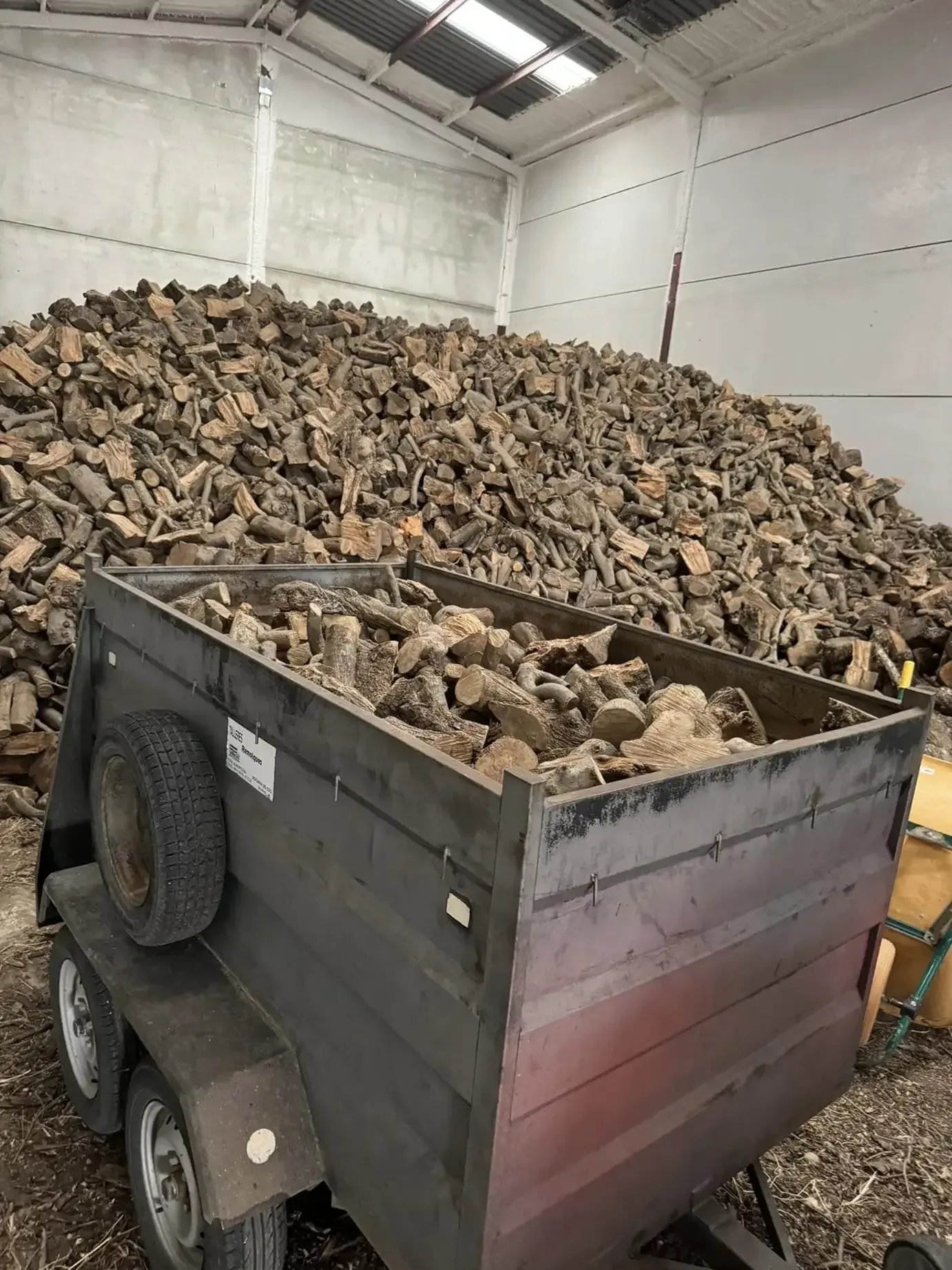 Un remolque lleno de trozos de madera está estacionado frente a una gran pila de madera dentro de un almacén.