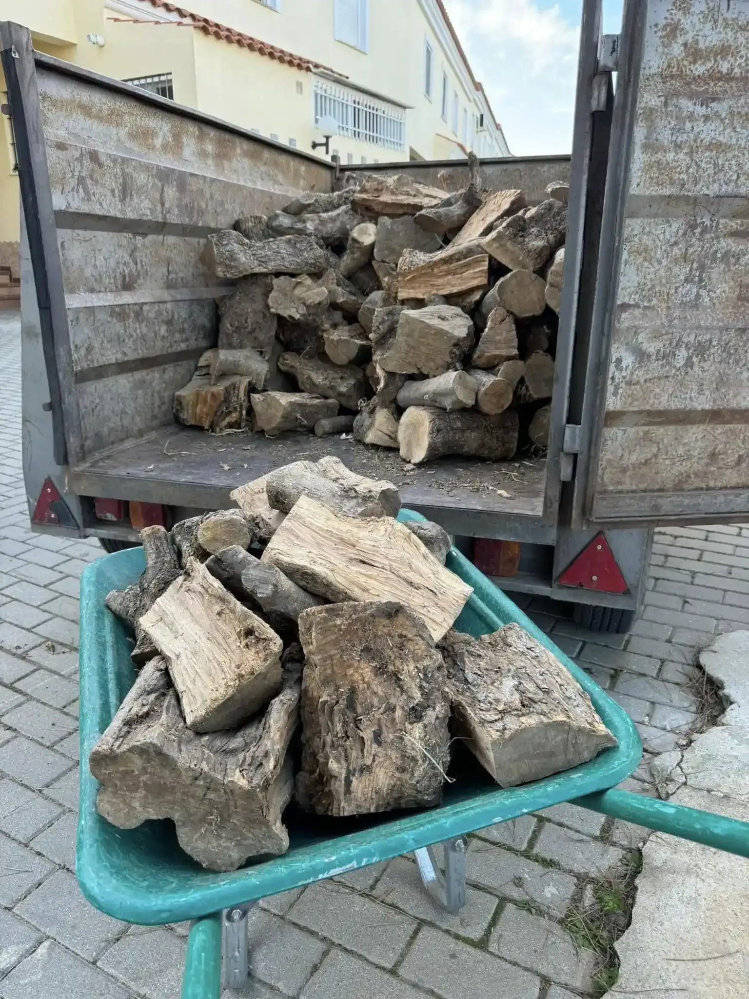 Carretilla llena de leña delante de un remolque también lleno de leña.