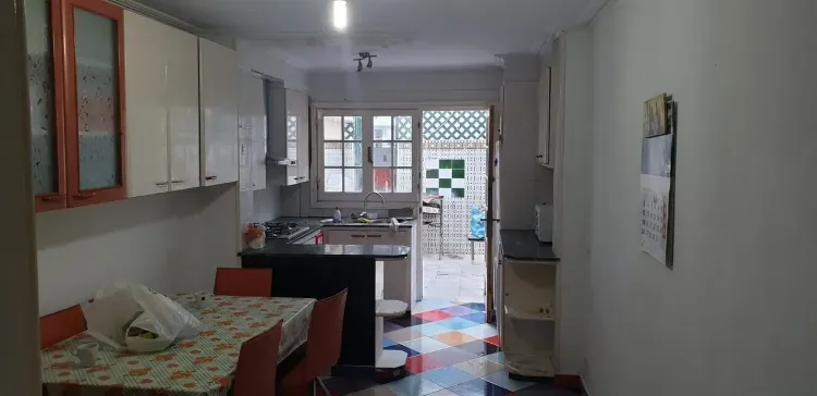 Cocina estrecha con armarios blancos, mesa y suelo de baldosas que da a una puerta luminosa.